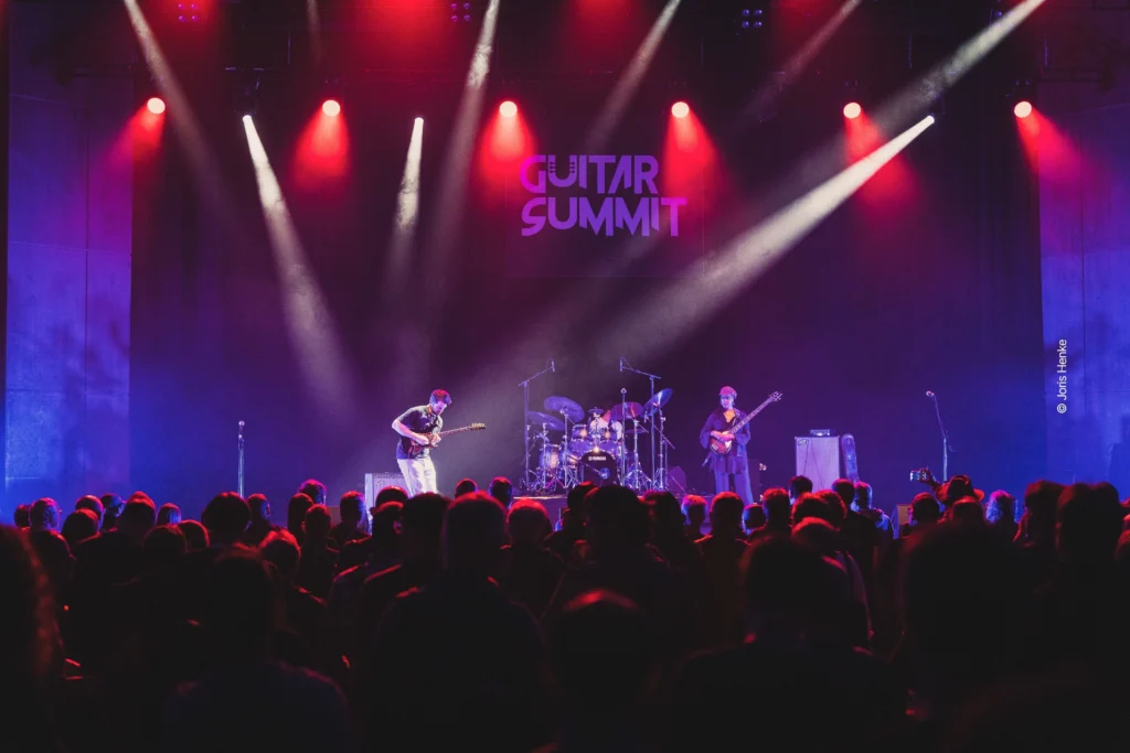 Dreiköpfige Band mit zwei Gitarren und einem Schlagzeug spielt auf beleuchteter Bühne in abgedunkelter Konzerthalle vor Publikum