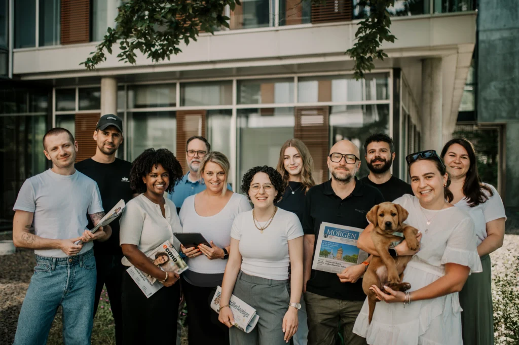 Gruppe von elf Mitarbeitenden der Haas Mediengruppe vor einem Gebäude, einige halten Zeitschriften, eine Person hält einen Hund im Arm.