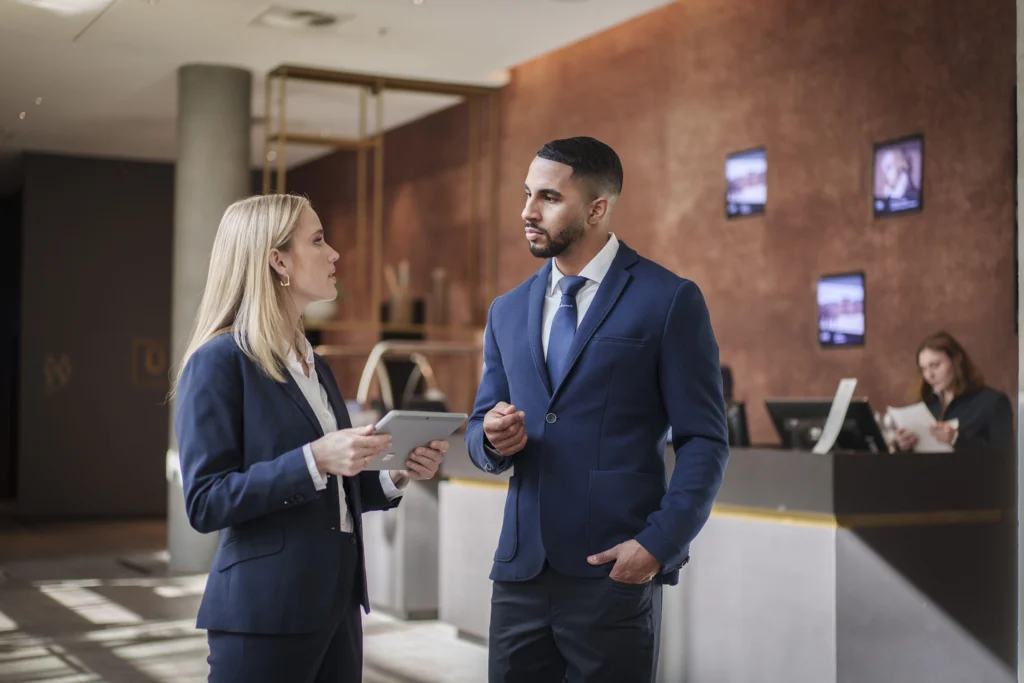 Zwei Personen in Businesskleidung unterhalten sich in einem modernen Hotellobby mit Rezeption im Hintergrund.