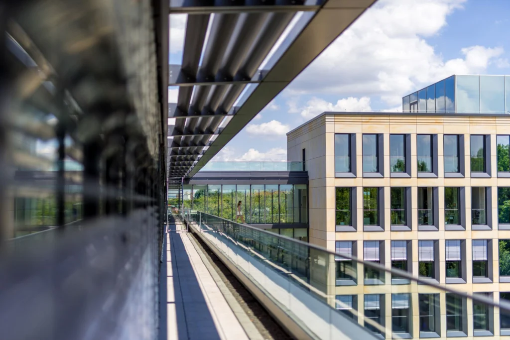 Blick entlang einer Glasbrüstung auf das moderne Bürogebäude der INTER Versicherung mit großen Fenstern und einem Vordach im Vordergrund, im Hintergrund blauer Himmel mit Wolken.