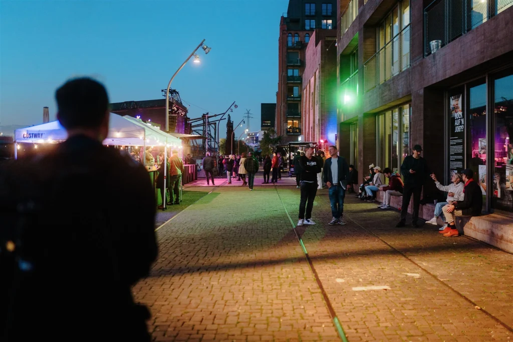 Abendliche Szene im Jungbusch mit beleuchteten Gebäuden, Gastronomie und Menschen auf der Straße.