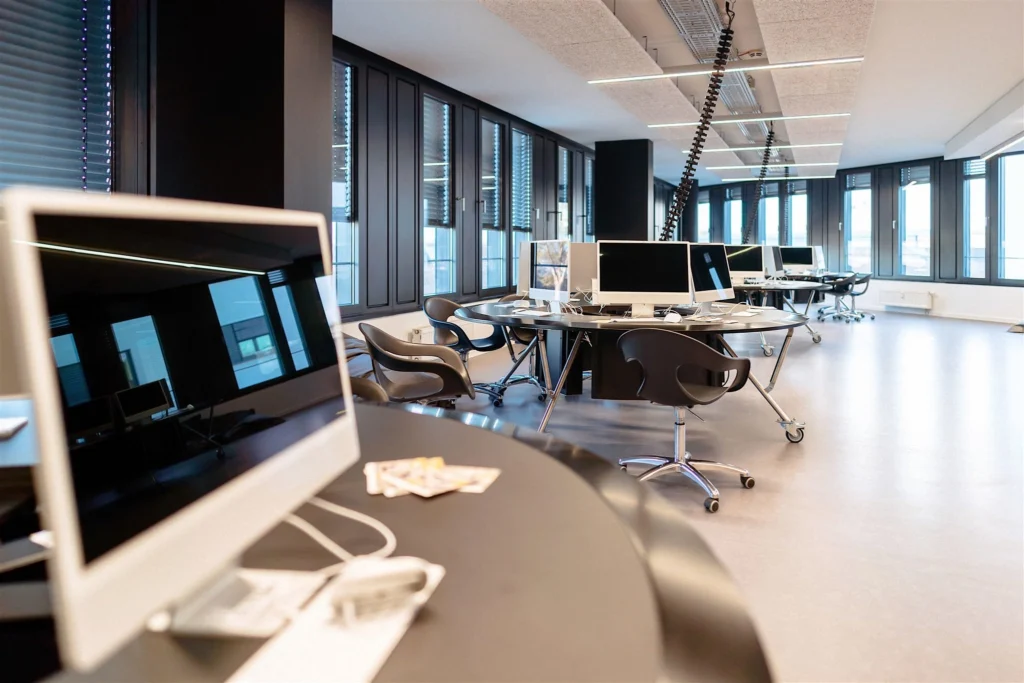 Moderner Büroraum mit mehreren Computern auf Tischen und ergonomischen Stühlen vor großen Fenstern.