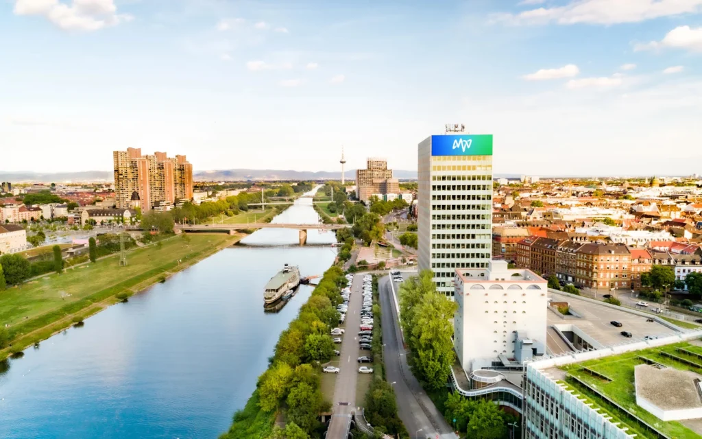 Luftaufnahme des MVV-Gebäudes in Mannheim neben dem Neckar mit Brücken, einem Schiff auf dem Fluss und umliegenden Wohn- und Bürogebäuden.