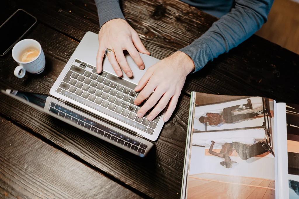Hände tippen auf einer Laptop-Tastatur auf einem dunklen Holztisch neben einer Tasse Kaffee und einem aufgeschlagenen Magazin mit Fotos von Menschen.