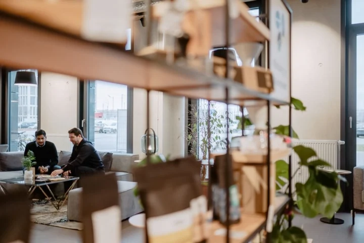 Two men sit at a low table in a modern, bright room with large windows and plants, with blurred shelves in the foreground.
