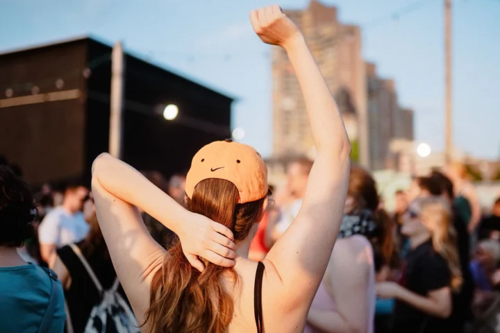 Person mit orangefarbener Kappe und langem Haar, die den Arm hebt und sich mit der anderen Hand am Nacken fasst, vor einer Menschenmenge am ALTER Mannheim.