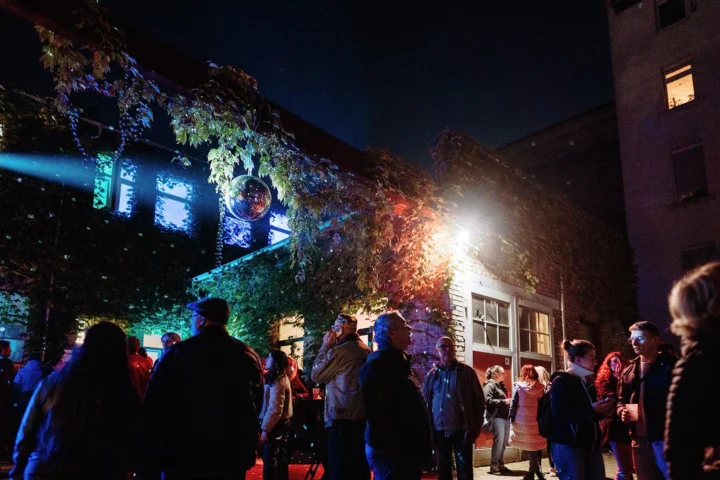 Besucherinnen und Besucher stehen am Abend vor einem begrünten Gebäude mit Disco-Kugel und farbiger Beleuchtung und unterhalten sich während einer Veranstaltung.