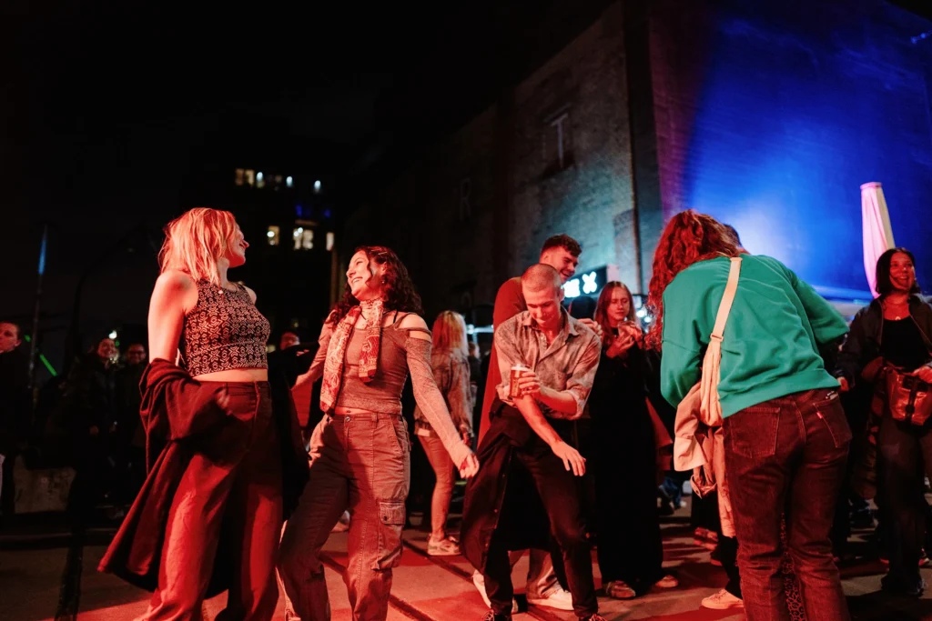 Mehrere Menschen tanzen und unterhalten sich bei einer Open-Air-Veranstaltung am Abend vor einer beleuchteten Bühne.