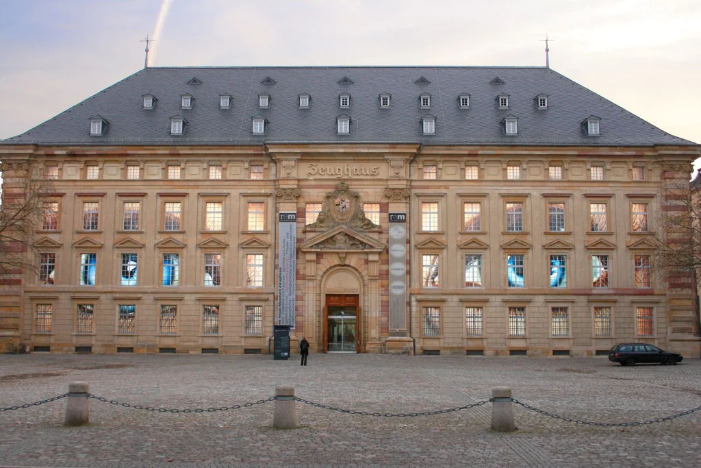 Das barocke Gebäude des Zeughauses in Mannheim mit symmetrischer Fassade, zahlreichen Fenstern, einem zentralen Eingang mit dekorativem Giebel und zwei vertikalen Bannern neben der Tür.