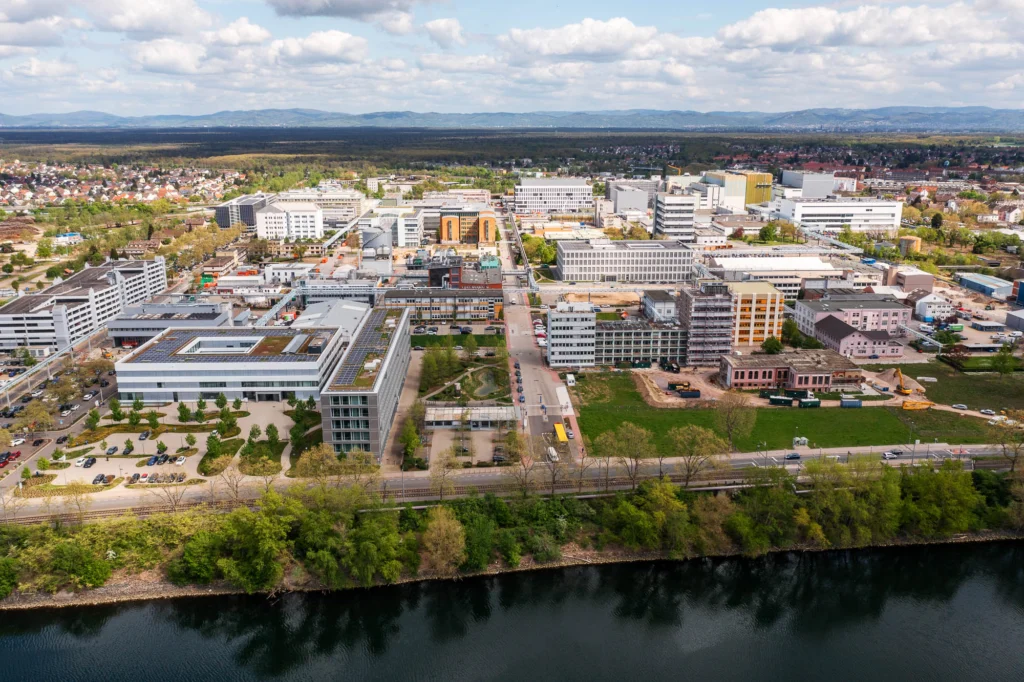 Luftaufnahme des Roche Firmengeländes in Mannheim mit mehreren modernen Büro- und Laborgebäuden entlang eines Flusses und einer Stadt im Hintergrund.