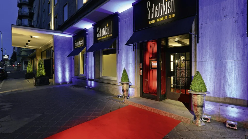 Eingangsbereich des Theaters Schatzkistl mit rotem Teppich, zwei großen Pflanzgefäßen und beleuchteter Fassade am Abend.