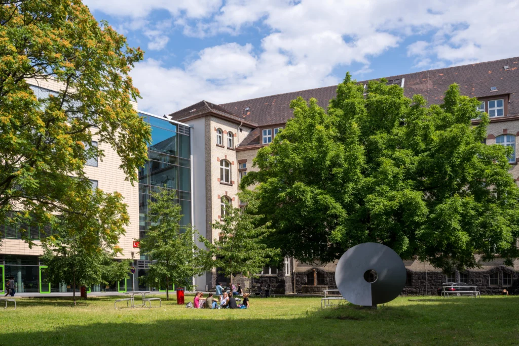 Campus mit Rasenfläche, Bäumen, einer großen runden Skulptur und mehreren Personen vor einem mehrstöckigen Gebäude mit Glasfassade und Ziegeldach.
