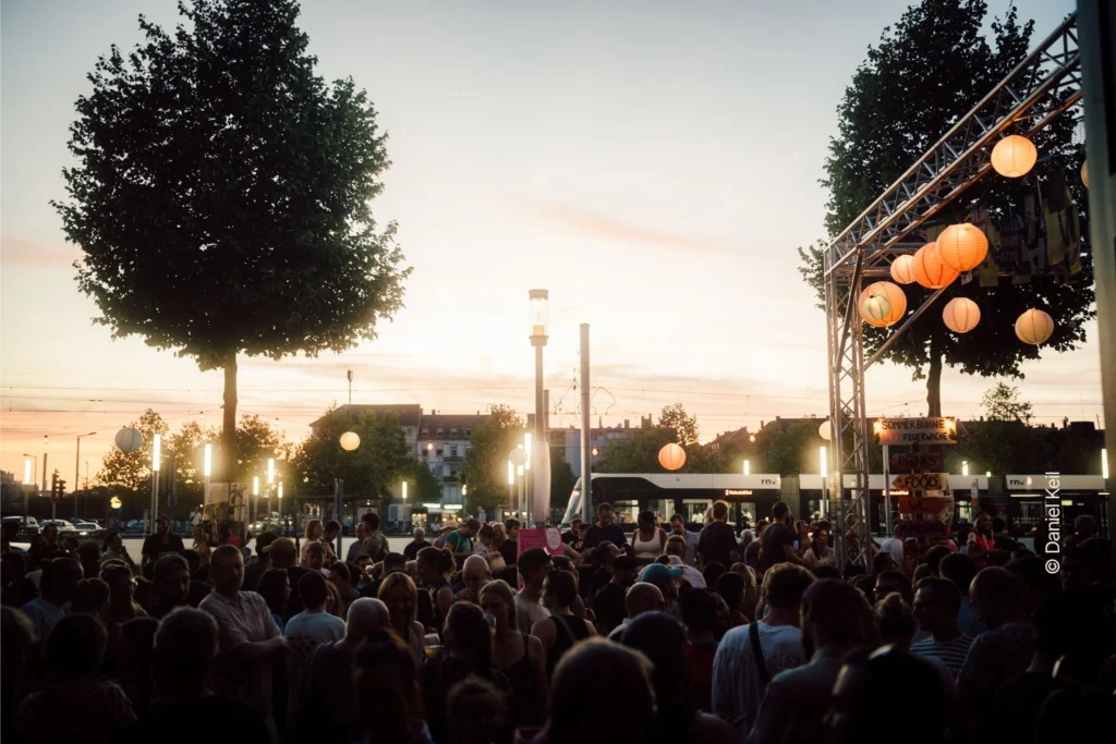 Menschen besuchen bei Sonnenuntergang eine gut besuchte Open-Air-Veranstaltung auf einem Platz mit beleuchteten Lampions, Bäumen und einer Straßenbahn im Hintergrund.