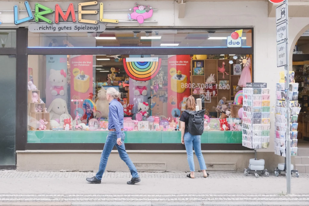 Die Schaufensterfront eines Spielwarengeschäfts mit dem Schriftzug „Urmel“ zeigt eine farbenfrohe Auslage, während Passantinnen und Passanten davor vorbeigehen.