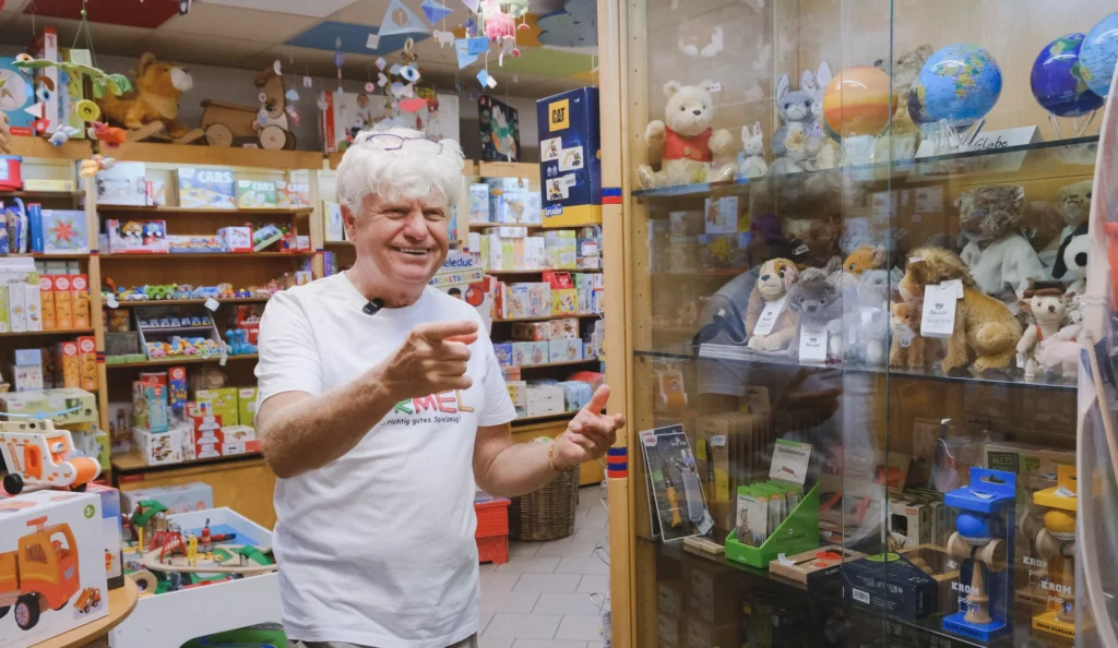 Eine Person steht in einem Spielwarengeschäft neben einer Glasvitrine mit Kuscheltieren und weiteren Spielwaren, während zahlreiche Produkte die Regale im Hintergrund füllen.