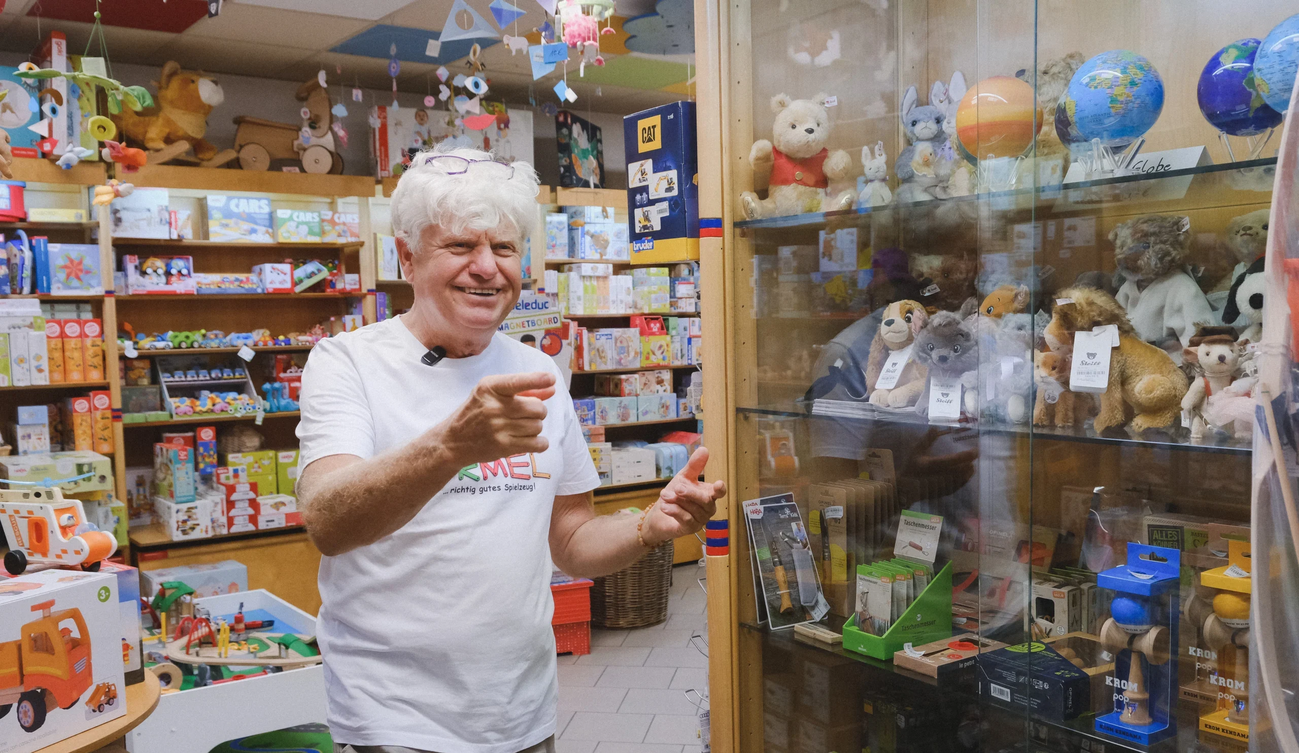 Eine Person steht in einem Spielwarengeschäft neben einer Glasvitrine mit Kuscheltieren und weiteren Spielwaren, während zahlreiche Produkte die Regale im Hintergrund füllen.