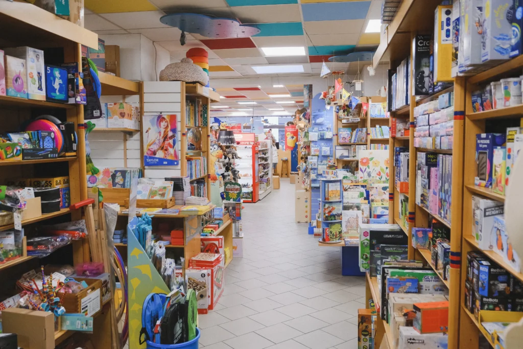 Der Blick durch einen Gang des Spielwarengeschäfts zeigt zahlreiche Regale mit Büchern, Spielen, Kreativmaterialien und Spielzeug unter einer farbig gestalteten Decke.