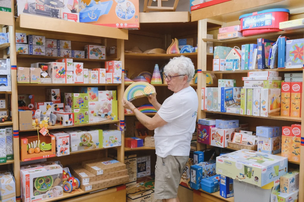Eine Person betrachtet einen regenbogenförmigen Holzbausatz vor Regalen mit pädagogischen Spielen, Baukästen und Holzspielzeug.