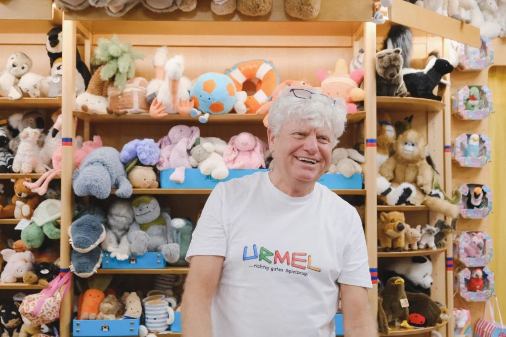 Eine Person mit weißem T-Shirt steht vor einem Regal voller Kuscheltiere und Spielwaren in einem bunt eingerichteten Spielwarengeschäft.
