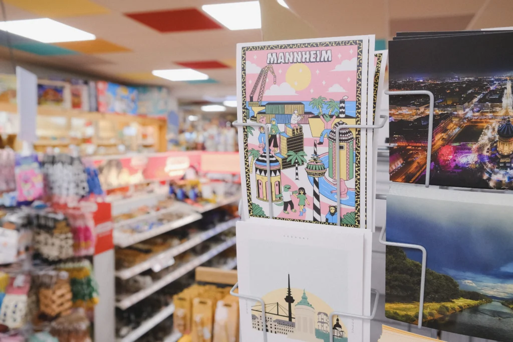 In einem Geschäft für Spielwaren und Souvenirs steht ein Drehständer mit farbenfrohen Mannheim-Postkarten und Stadtmotiven vor unscharfen Verkaufsregalen im Hintergrund.