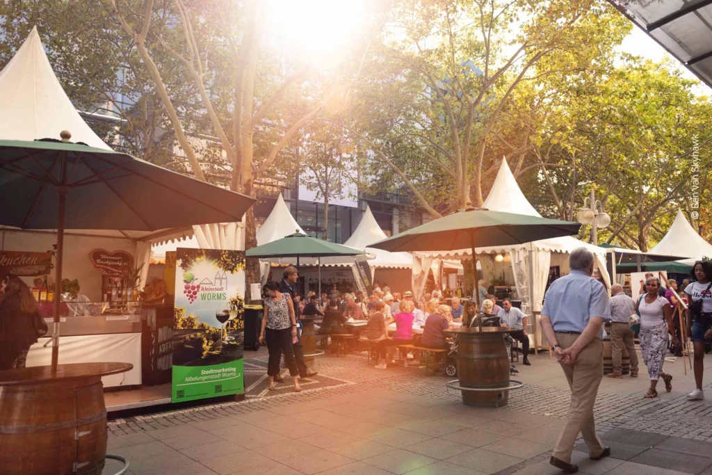 Marktstände mit Sonnenschirmen und Zelten auf den Kapuzinerplanken mit Bäumen im Hintergrund. Menschen sitzen an Tischen und laufen über das Fest.