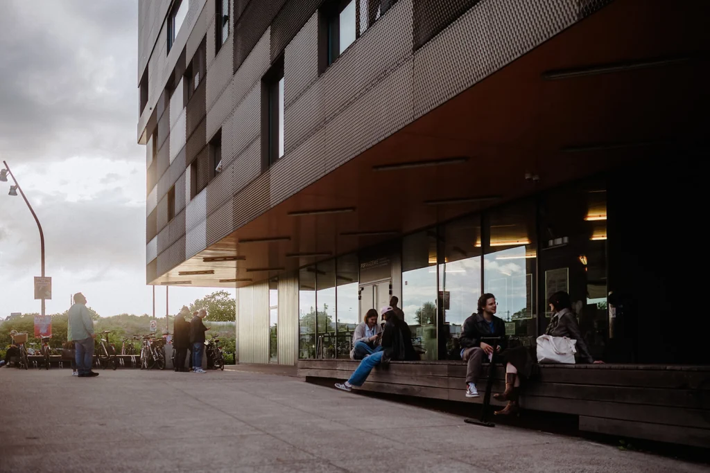 Menschen sitzen und stehen vor der Popakademie mit großer Glasfront, Fahrräder sind daneben abgestellt, abends bei warmem Licht.