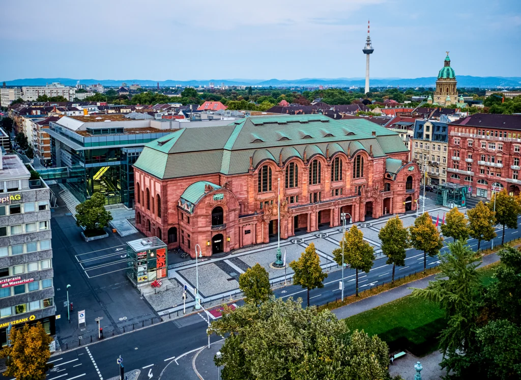 Luftaufnahme des historischen Rosengarten-Gebäudes in Mannheim mit umliegenden Straßen, Bäumen und dem Fernsehturm im Hintergrund.