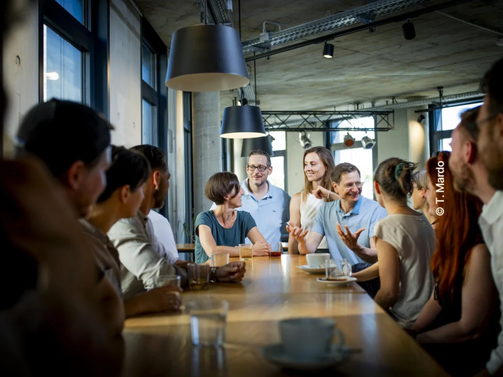 Menschen sitzen an einem langen Tisch mit Getränken und unterhalten sich.
