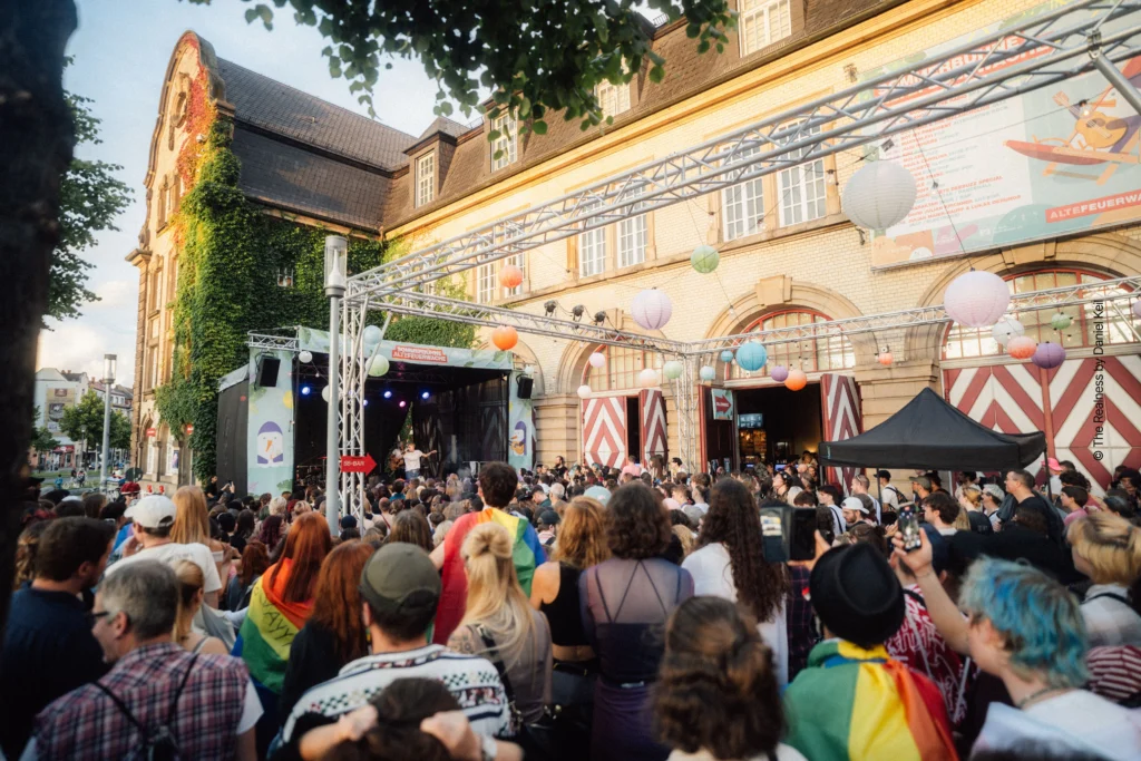 Menschen besuchen eine gut besuchte Open-Air-Veranstaltung auf einem Platz mit Lampions, Bäumen und der Alten Feuerwache im Hintergrund