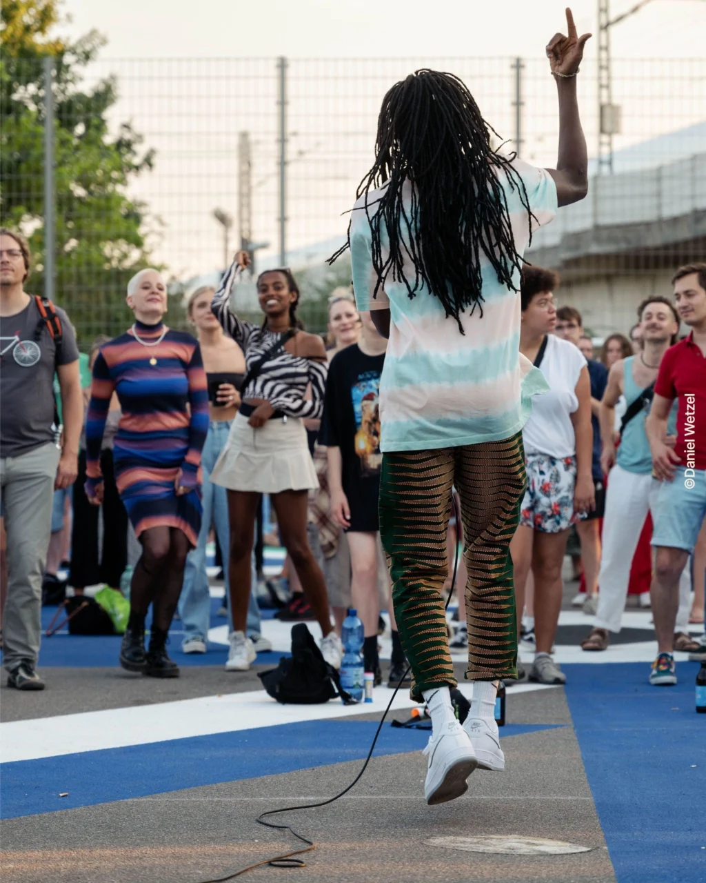 Person mit langen Dreadlocks springt mit erhobenem Arm vor einer Gruppe von Menschen auf einem urbanen Platz.